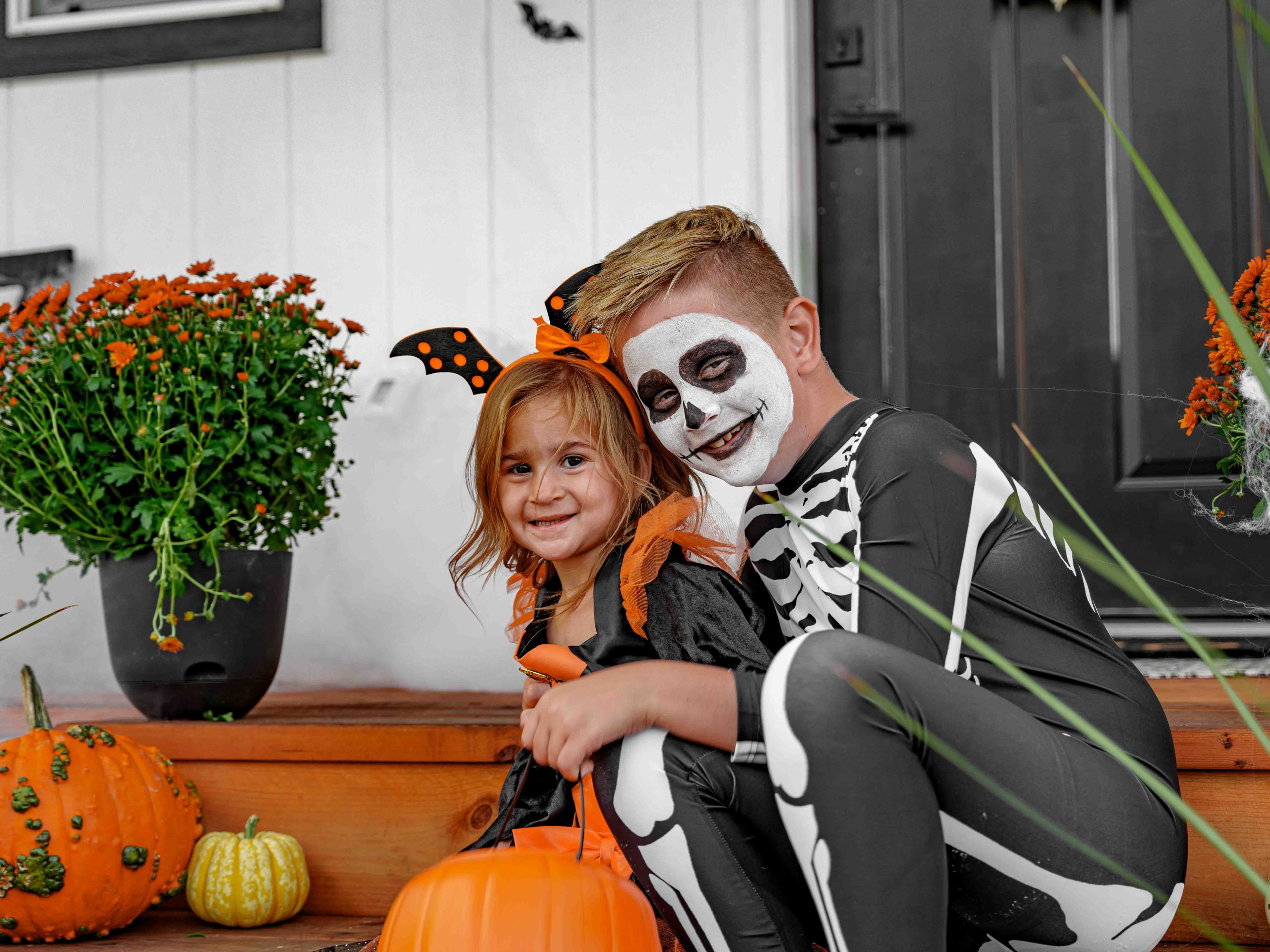 Children in bright orange and black costumes laughing together on a front porch while one holds a small chalkboard with a joke written on it
